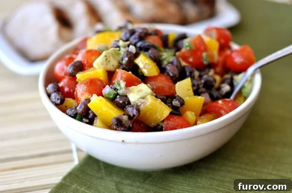 A beautifully presented white bowl filled with a fresh black bean and veggie guacamole salad, garnished with cilantro and a lime wedge.