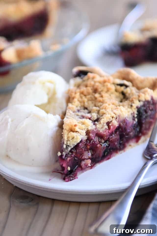 A beautiful Triple Berry Rhubarb Pie slice elegantly presented on a white plate with a scoop of vanilla ice cream, ready to be savored.