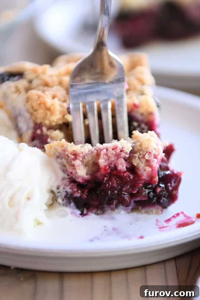 A fork digging into a slice of triple berry rhubarb pie, accompanied by a melting scoop of vanilla ice cream.