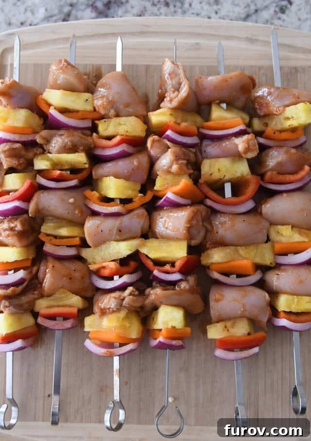 Aloha Chicken Pineapple Kabobs 4 Six vibrant metal skewers laden with perfectly arranged pieces of chicken, golden pineapple, colorful bell peppers, and red onions. They are neatly placed on a rustic wood cutting board, ready for grilling or serving.