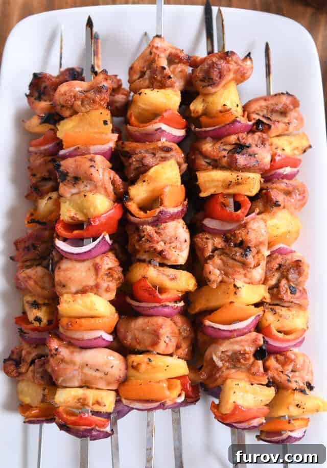 Aloha Chicken Pineapple Kabobs 5 Five gleaming metal skewers loaded with marinated chicken pieces, bright pineapple chunks, vibrant bell peppers, and red onions, sizzling on a hot grill, showing delicious char marks.