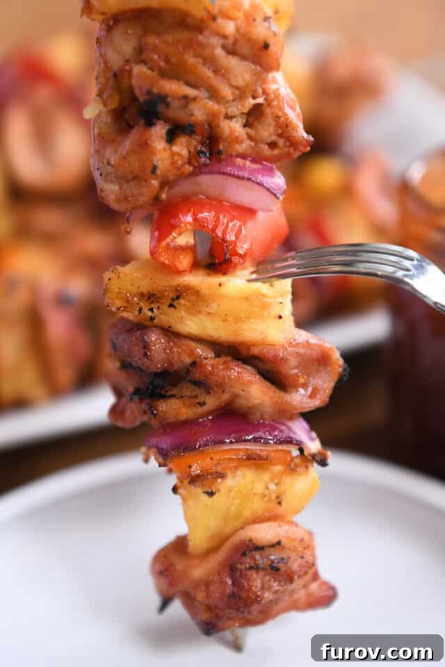 Aloha Chicken Pineapple Kabobs 6 A fork gently presses down on a piece of golden-brown grilled pineapple, highlighting its caramelized perfection on a metal skewer, alongside tender chicken, onions, and peppers.