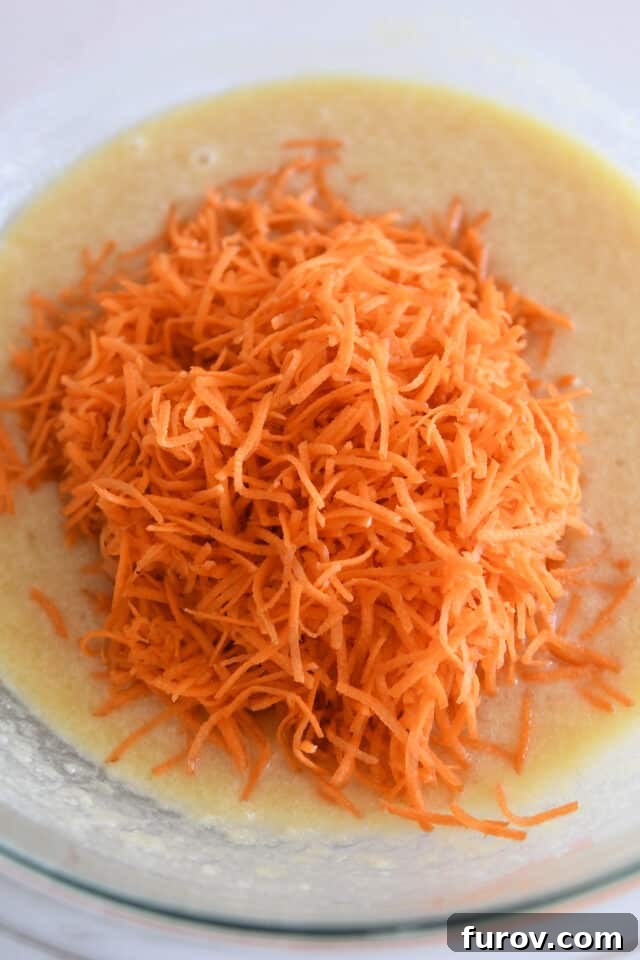 Finely shredded carrots being added to the glass bowl with cake batter, ensuring moisture and flavor throughout.