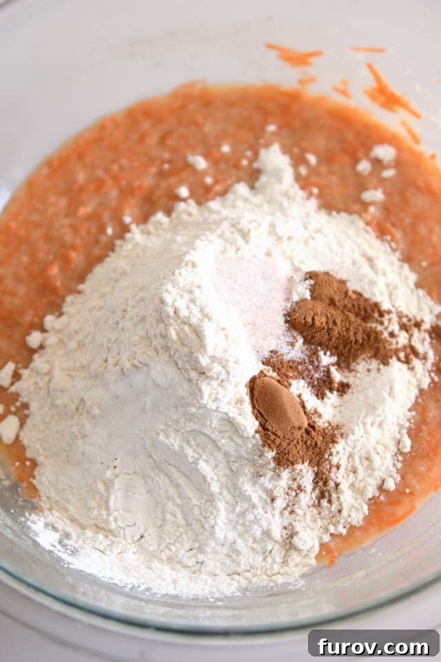 Flour, cinnamon, salt, and baking soda being mixed into the carrot cake batter in a glass bowl, dry ingredients completing the mix for a perfect rise.