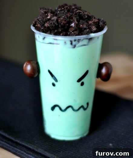 A clear plastic cup with bright green pudding, topped with dark crushed Oreo cookies. A playful monster face is drawn on the outside of the cup with a black marker, featuring wide eyes and a smiling mouth.