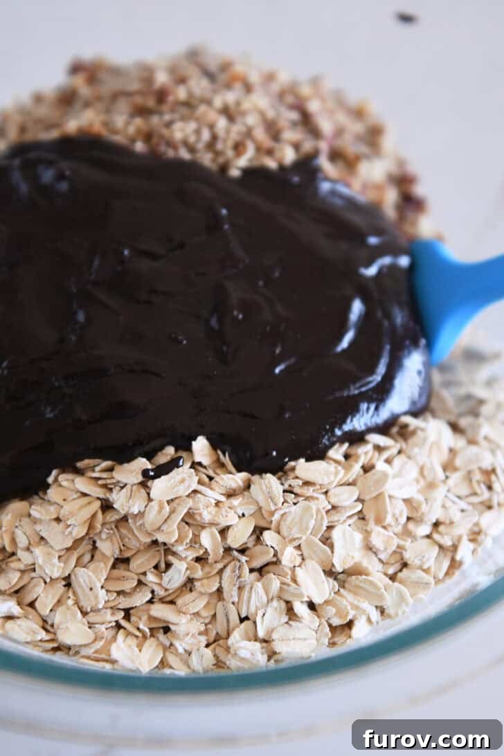 Thick chocolate syrup mixture being poured over a bowl of rolled oats and chopped pecans, ready to be mixed for homemade granola.