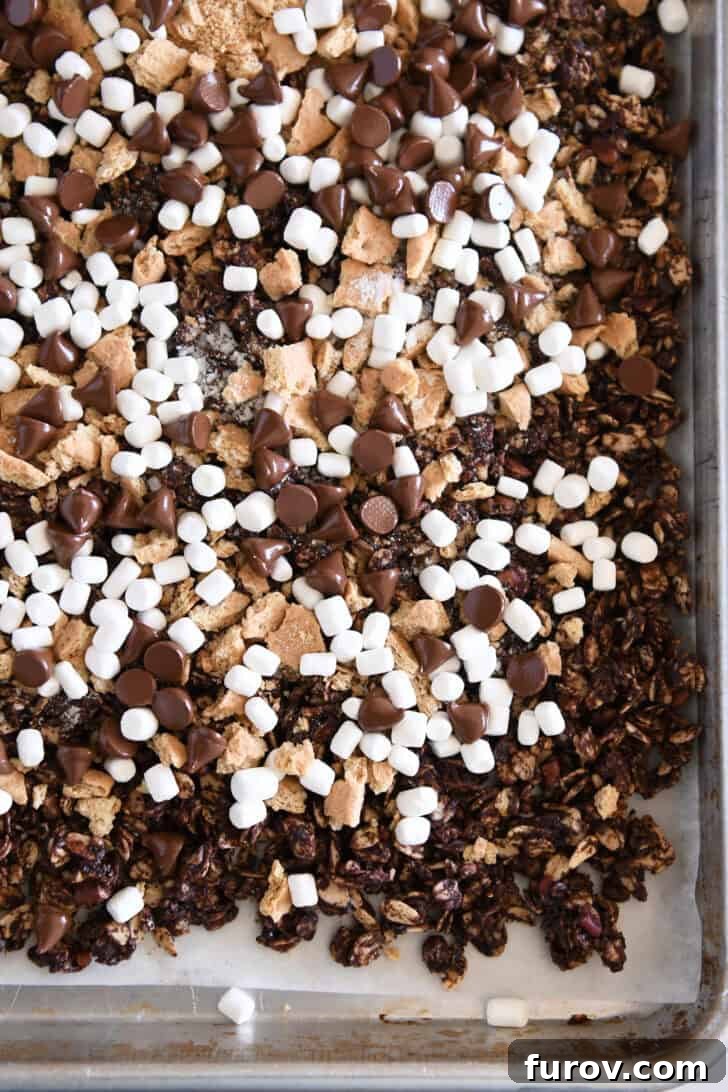 Freshly baked chocolate s'mores granola on a parchment-lined sheet pan, generously topped with golden graham cracker pieces, fluffy mini marshmallows, and glistening melty chocolate chips.