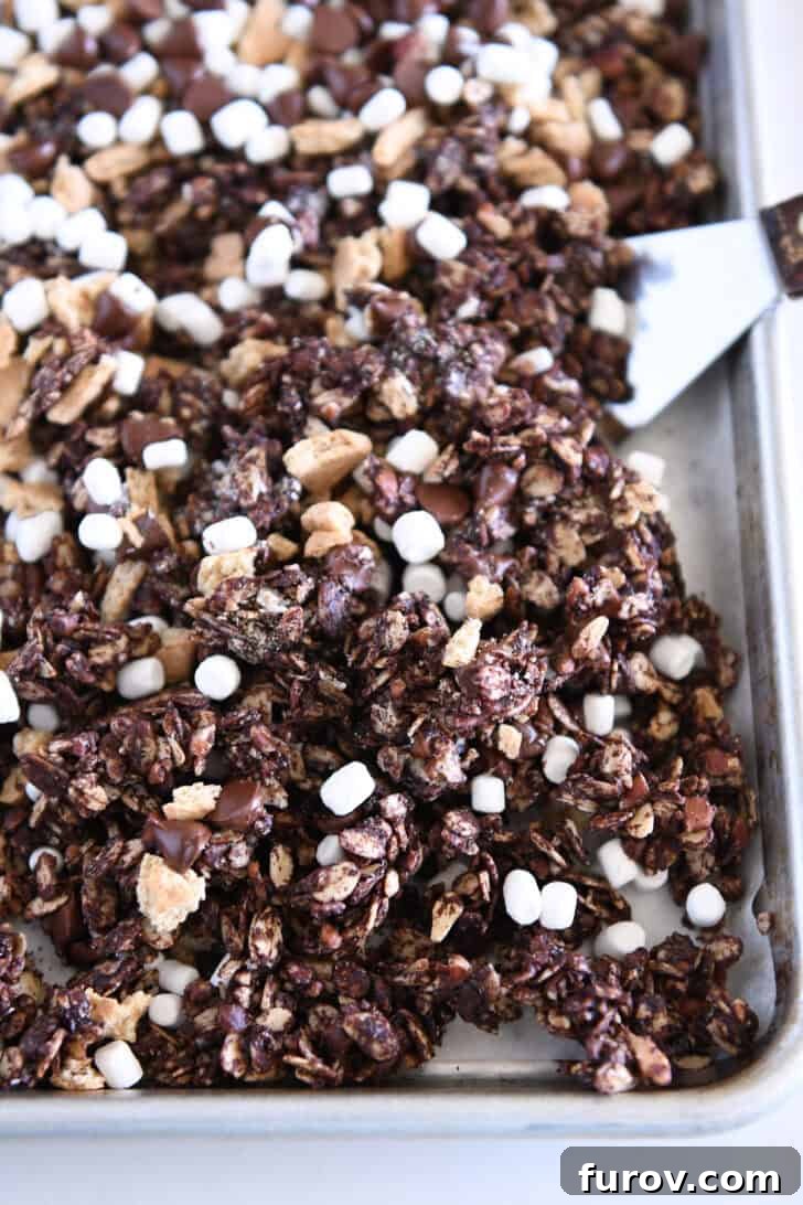A close-up shot of chocolate granola spread on a parchment-lined sheet pan, adorned with chunky graham cracker pieces, mini marshmallow bits, and dark chocolate chips, ready for its final bake.