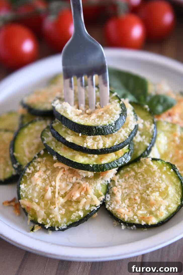 Fork spearing three golden-brown zucchini rounds on a gray plate, showcasing the crispy Parmesan topping.