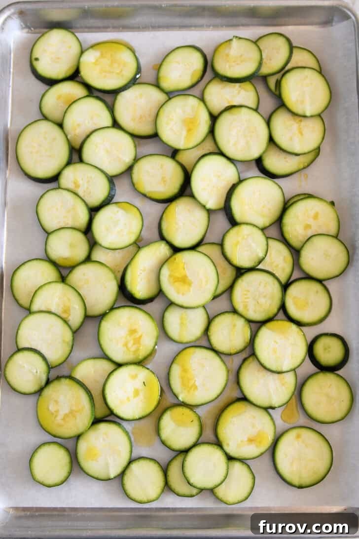 Thinly sliced zucchini rounds arranged on a parchment-lined half sheet pan, drizzled with olive oil.