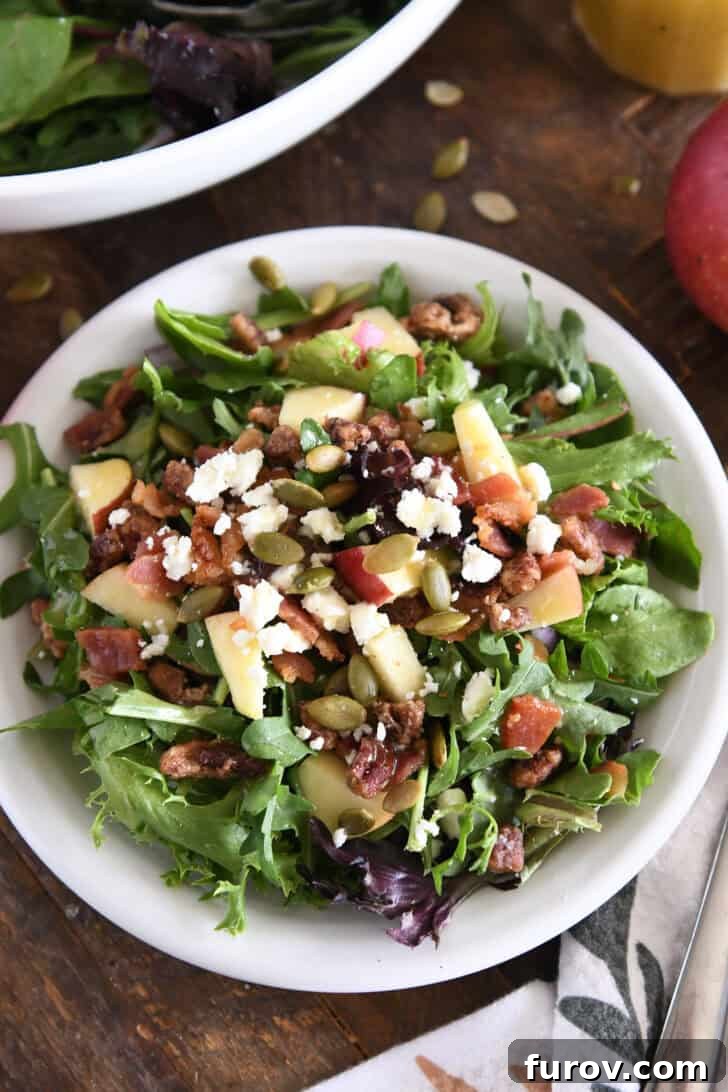A beautifully arranged harvest apple salad with roasted pumpkin seeds, chopped apples, candied pecans, crumbled feta cheese, and thinly sliced red onions on a white plate, viewed from above.