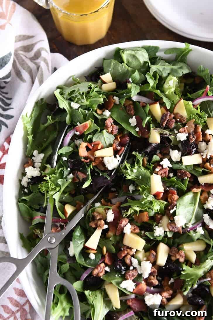 A white bowl brimming with a vibrant mixed salad, showcasing baby greens, chunks of crisp apples, creamy feta cheese, savory cooked bacon bits, sweet dried cranberries (craisins), and roasted pumpkin seeds. Salad tongs rest in the bowl, inviting a generous serving.
