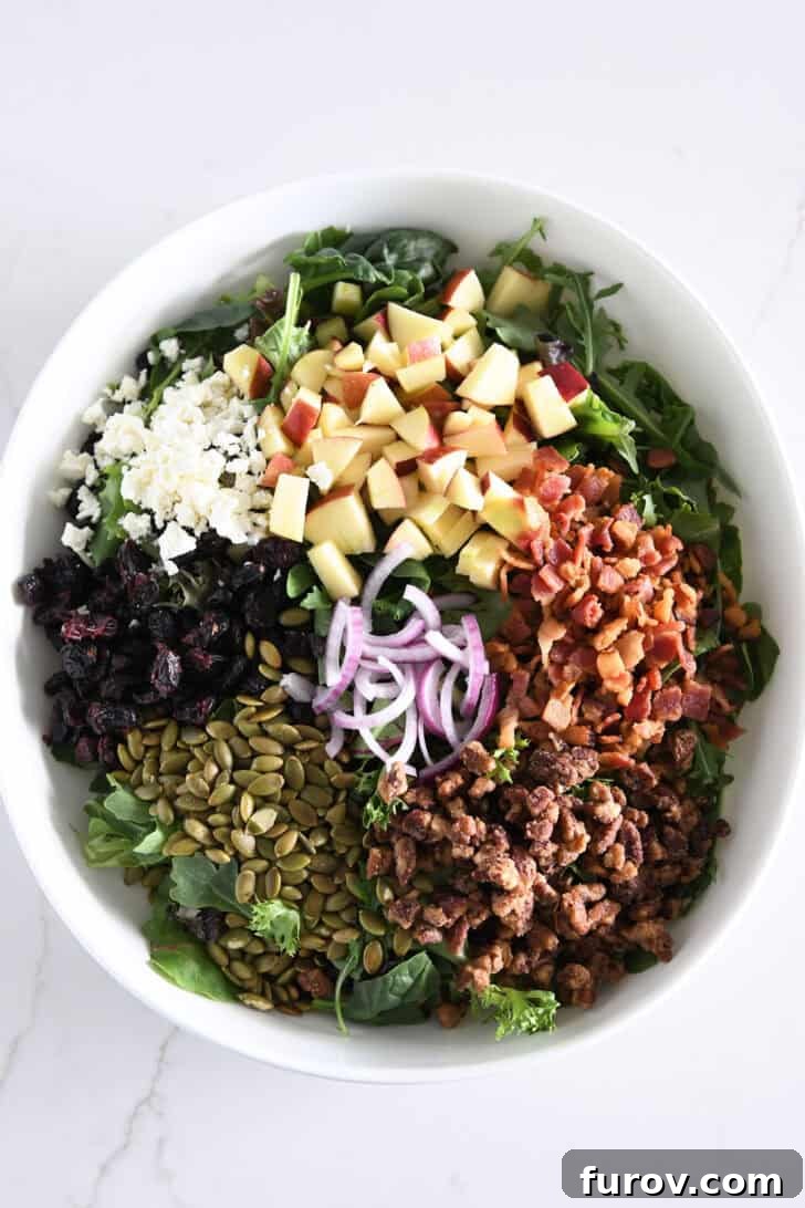 A white bowl showcasing the fully assembled Harvest Apple Salad with crisp green leaves, roasted pumpkin seeds, sweet candied pecans, crumbled cooked bacon, vibrant red onions, dried cranberries, creamy feta, and freshly chopped apples.