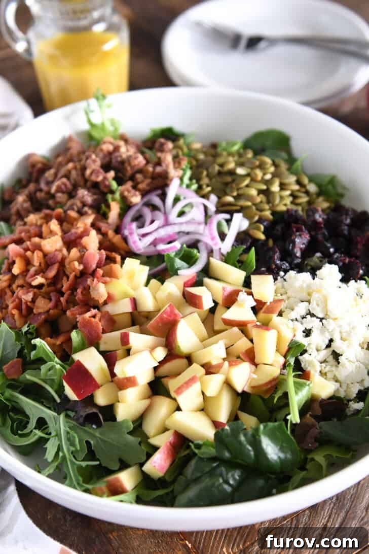 A close-up view of a selection of Harvest Apple Salad ingredients: bright red chopped apples in the foreground, with creamy feta cheese, crispy bacon bits, sweet candied pecans, earthy roasted pumpkin seeds, and thinly sliced red onions artfully arranged in the background.