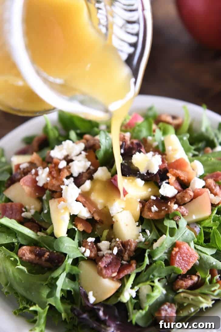 A stream of vibrant apple cider vinaigrette being generously poured over a fresh green salad, which includes crisp chopped apples, creamy feta cheese, crunchy pecans, and savory bacon pieces, glistening under the dressing.