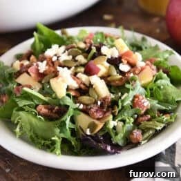 A vibrant green salad on a white plate, garnished with roasted pumpkin seeds, crisp chopped apples, sweet candied pecans, crumbled feta cheese, and thinly sliced red onions.
