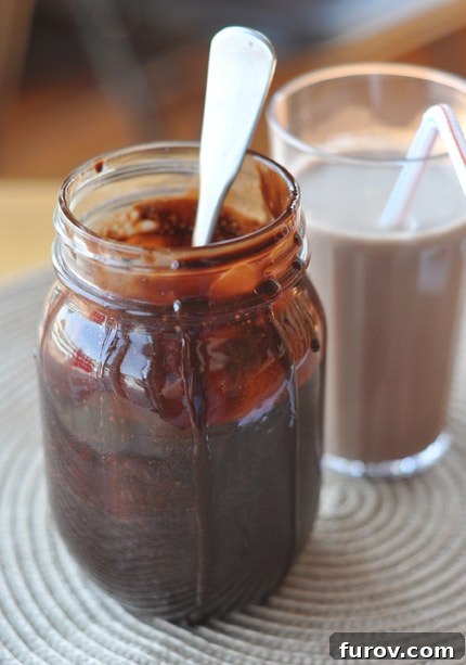 A charming mason jar filled with rich, homemade chocolate syrup, with delightful drips running down the side, positioned invitingly in front of a perfectly mixed glass of chocolate milk.