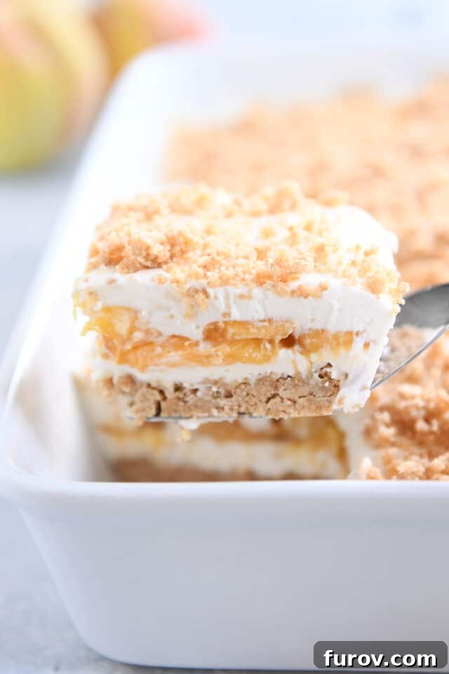 No-Bake Peaches and Cream Bliss 2 A square slice of no-bake peaches and cream dessert rests on a metal spatula, held over a white 9x13-inch pan. The dessert shows distinct layers of golden crust, creamy white filling, and vibrant peach slices.