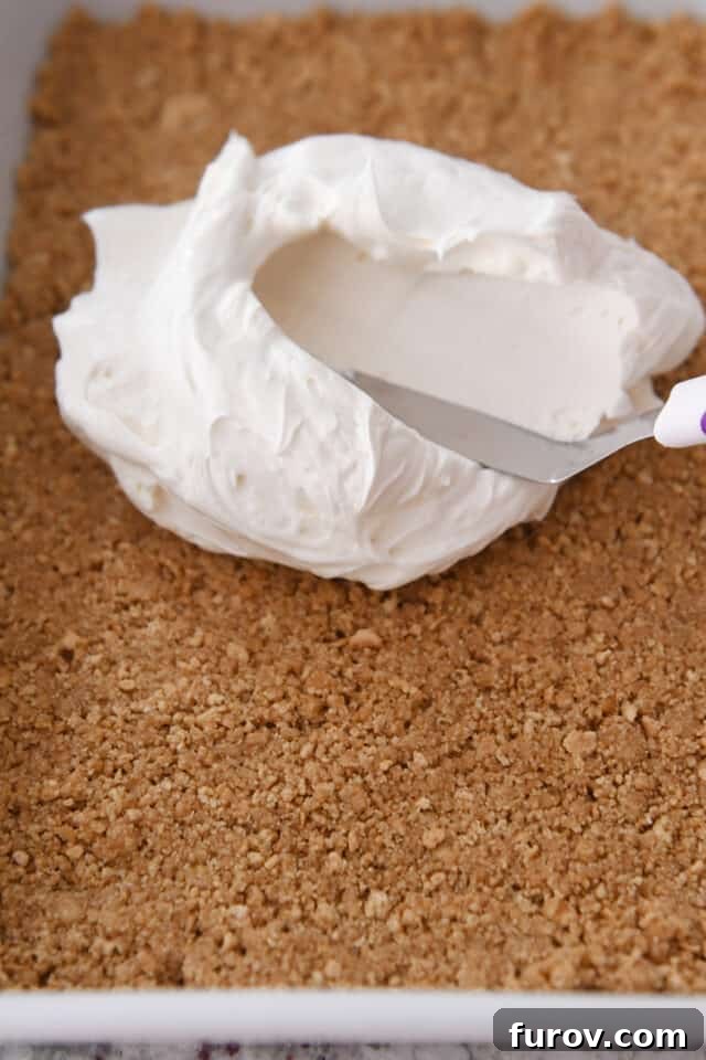 No-Bake Peaches and Cream Bliss 4 Hands carefully spreading a thick, luxurious cream cheese filling over a golden graham cracker crust in a baking pan, showing the start of dessert assembly.