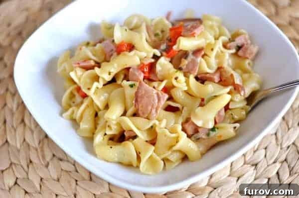 Creamy pasta with diced ham and red peppers in a white serving bowl, garnished with fresh parsley.