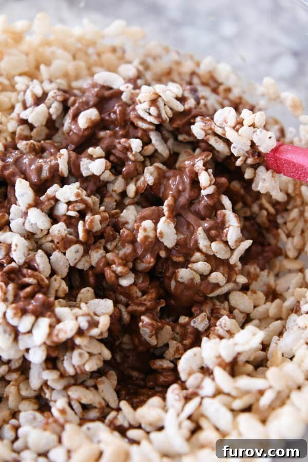 A spatula stirring crisp rice cereal into a rich peanut butter chocolate mixture in a bowl.
