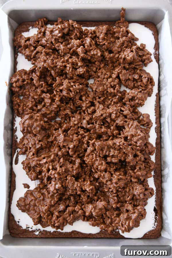 An offset spatula spreading the crispy peanut butter chocolate topping evenly over the marshmallow cream on a pan of brownies.