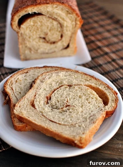 Golden Cinnamon Swirl Loaf 3 A freshly baked loaf of cinnamon swirl bread sits on a rustic cutting board, with two perfectly sliced pieces arranged on a white plate in front, showcasing the elegant spiral of cinnamon.