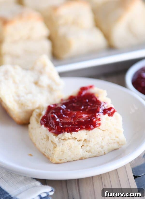 A super flaky buttermilk biscuit on a white plate, cut in half and served with dollops of sweet jam and creamy butter.