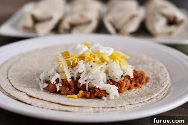A tortilla with beef, beans, white rice, cheese, and sour cream on it, ready to be rolled into a burrito.