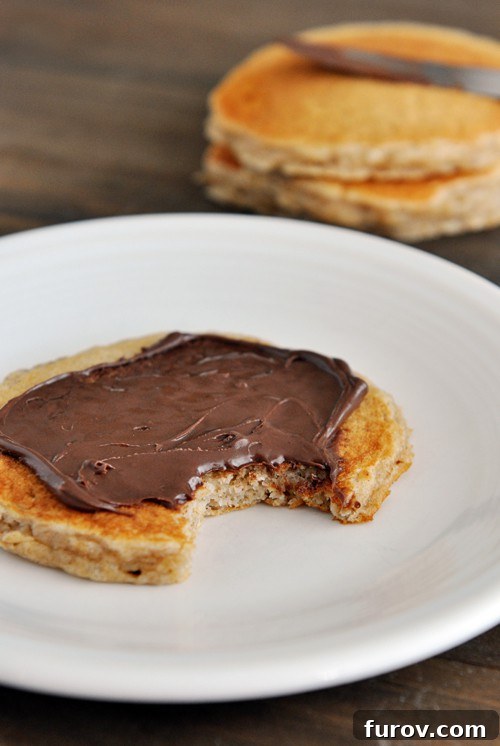 A pancake topped with Nutella, with a bite taken out, on a white plate.