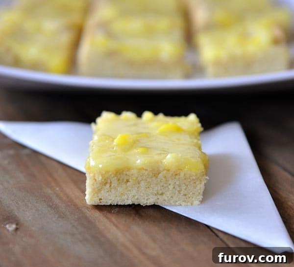 A perfectly sliced lemon bar, generously topped with a glistening lemon drop glaze and garnished with tiny lemon candies, resting in front of a tray of more freshly baked bars.