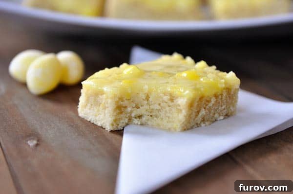A close-up of a Lemon Drop Sugar Cookie Bar with a delicate bite taken out, revealing the tender cookie interior beneath the shiny lemon glaze, with scattered lemon drops beside it on a white surface.