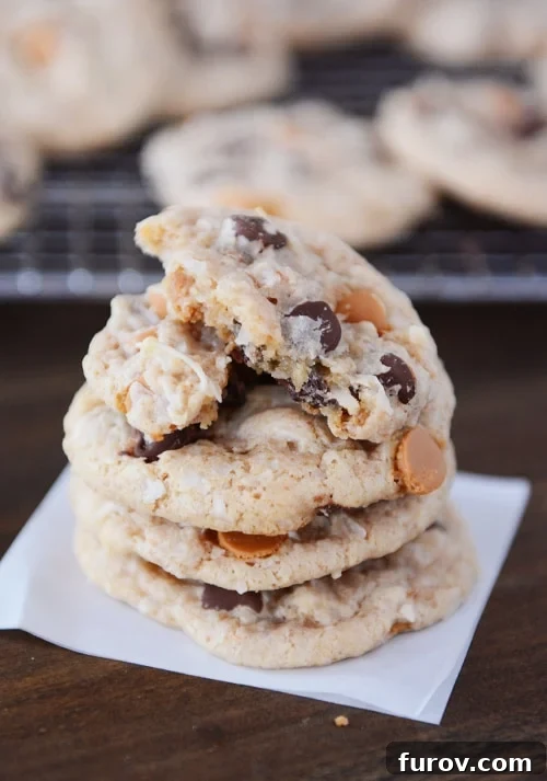 A stack of golden brown 7-Layer cookies featuring visible chocolate and butterscotch chips, with the top cookie split in half to reveal its soft, chewy interior.