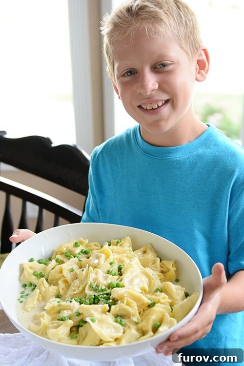 Snapshot Saturday: Mastering Mealtime Mayhem 2 A young boy proudly holding a white bowl filled with creamy tortellini Alfredo pasta and green peas, showcasing his culinary accomplishment.