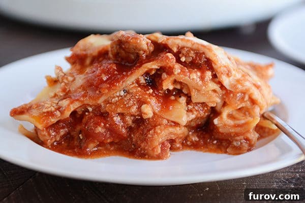 A white plate showcasing a generous slice of hot, cheesy slow cooker lasagna, topped with rich red sauce and melted mozzarella.