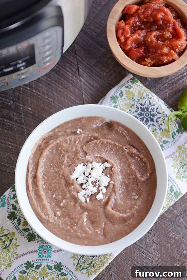 A white bowl of refried beans with a spoon on the side and cheese sprinkled in the middle of the bowl. There is a wooden bowl of chopped tomatoes in the background. 