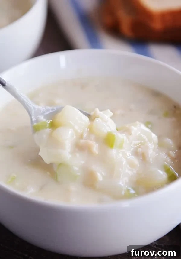 Our Go-To New England Clam Chowder 2 A spoon scooping a generous portion of creamy New England clam chowder from a white bowl, showing tender clams and potatoes.