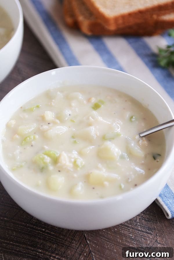 Our Go-To New England Clam Chowder 4 A pristine white bowl filled with creamy New England clam chowder, with a spoon resting in it, ready to be enjoyed.