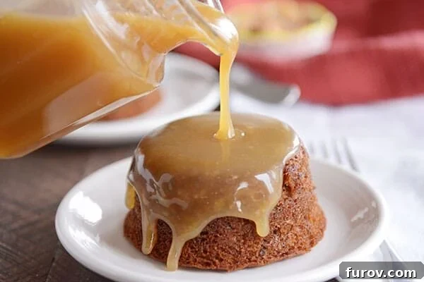 A small, brown cake on a white plate, with light brown sticky toffee sauce being poured over the top. 