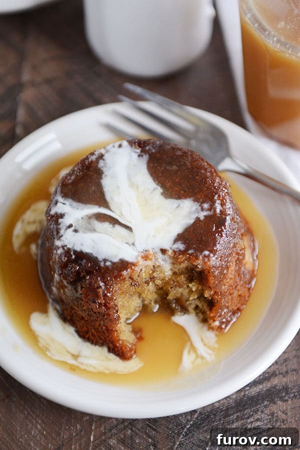 A small brown cake, with a bite taken out, covered with sticky toffee sauce and cream on a white plate.