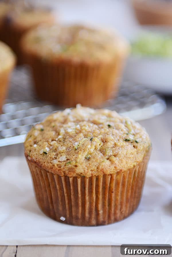 Ultimate Wholesome Zucchini Muffins 6 A perfectly baked zucchini muffin, still warm, resting on parchment paper with a backdrop of other muffins cooling on a rack, emphasizing its artisanal quality.