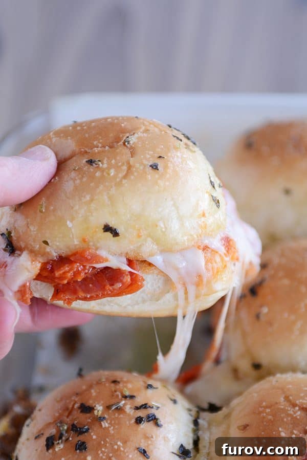 A hand reaching to pull apart a single cheesy pizza slider from a larger tray.
