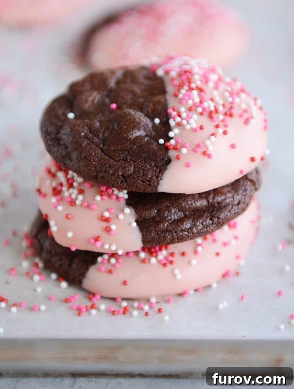 Decadent One-Bowl Fudgy Brownie Cookies 5 A charming stack of chocolate cookies, half-dipped in white frosting with rainbow sprinkles, presented on a rustic wooden surface.