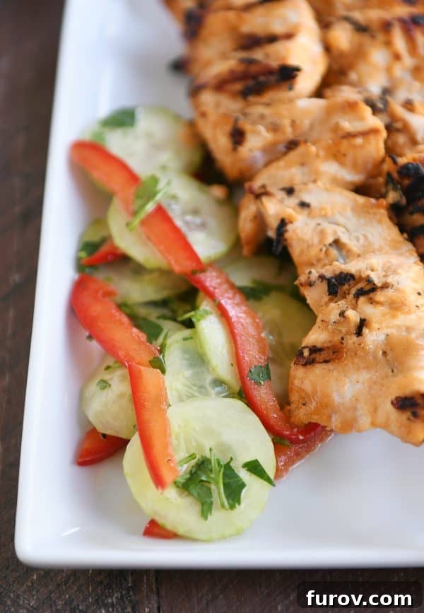 Close-up shot of grilled almond coconut milk chicken satay skewers and a fresh cucumber salad on a white plate, highlighting the textures and vibrant colors.
