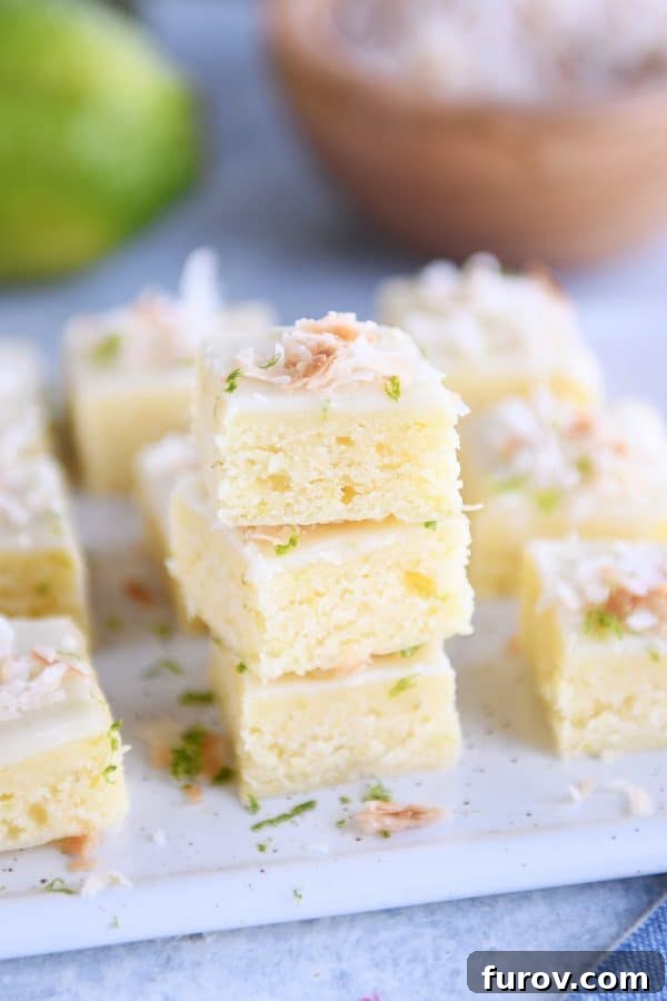 Several toasted coconut lime bars stacked artfully on a white platter, highlighting their thick glaze and coconut topping.