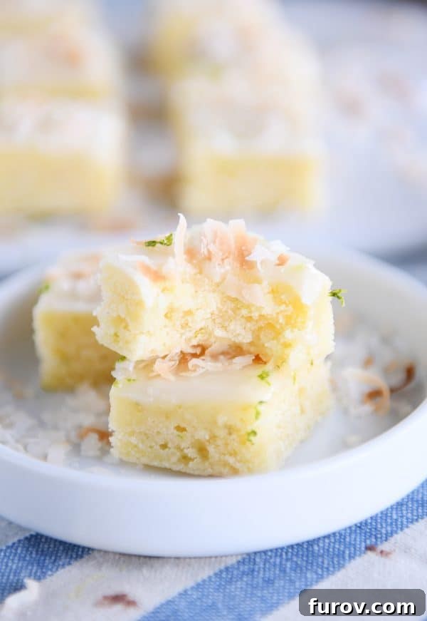 Close-up of a toasted coconut lime bar with a bite taken out, showing its moist interior and thick glaze, stacked on a small white plate.