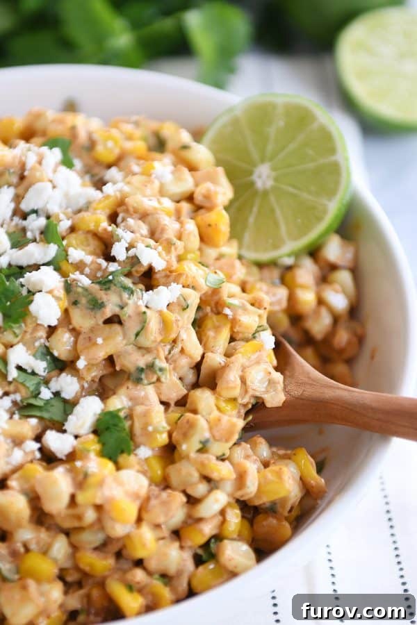 Zesty Mexican Street Corn Salad 2 A vibrant bowl of Mexican street corn salad, garnished with fresh chopped cilantro and lime wedges, with a wooden spoon ready for serving.