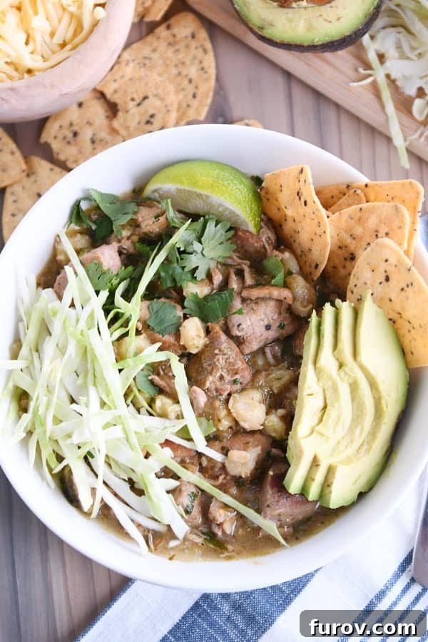 Top down view of slow cooker posole in white bowl with shredded cabbage, sliced avocado, tortilla chips and lime wedge, ready to be served.