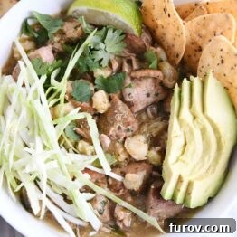 top down view of slow cooker posole in white bowl with shredded cabbage, sliced avocado, tortilla chips and lime wedge