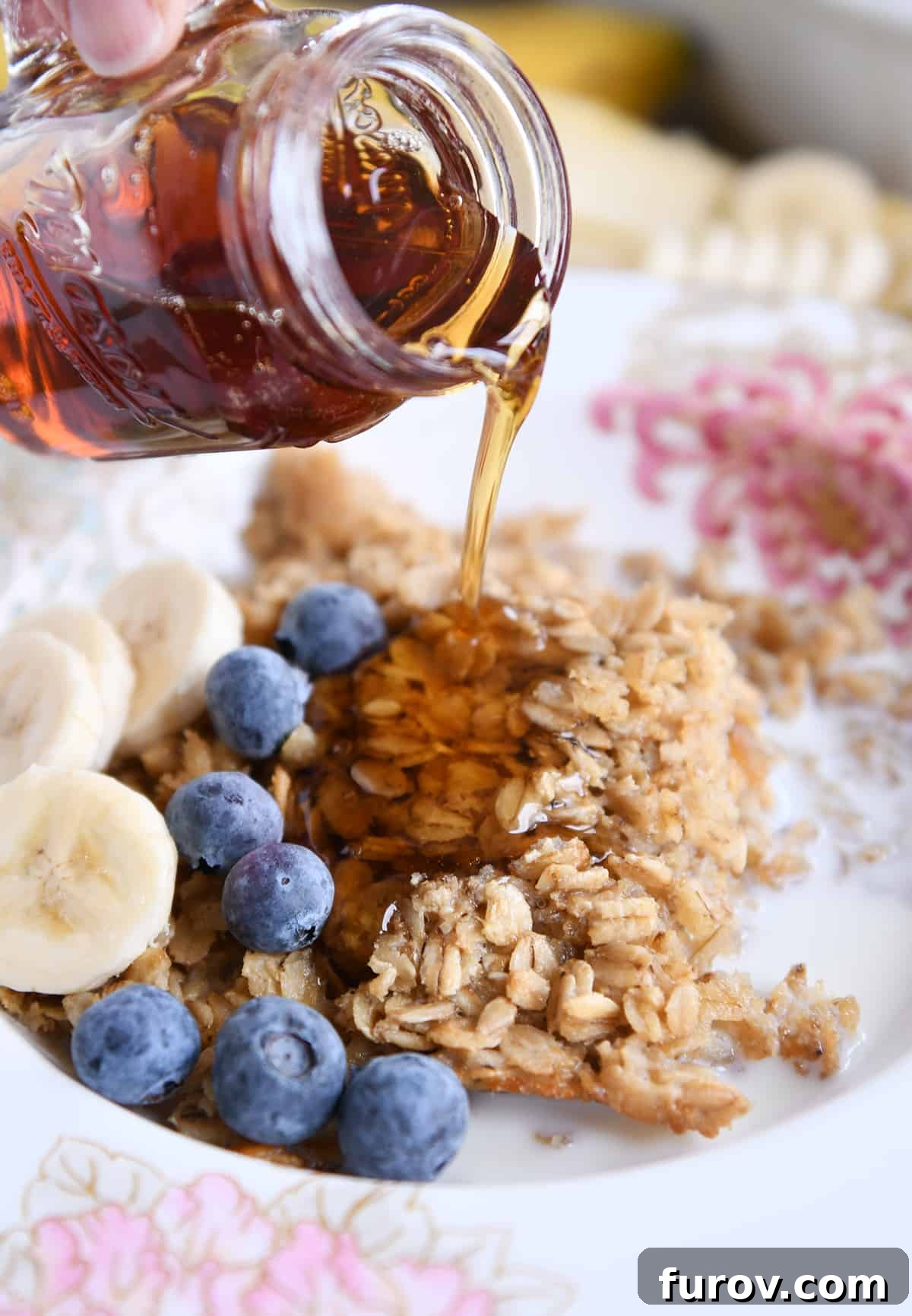 A stream of golden maple syrup being poured over a serving of Amish baked oatmeal, adorned with vibrant blueberries and banana slices.
